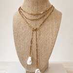 A gold-toned multi-strand necklace with two large white shell-like pendants, displayed on a beige fabric-covered jewelry bust against a light neutral background.