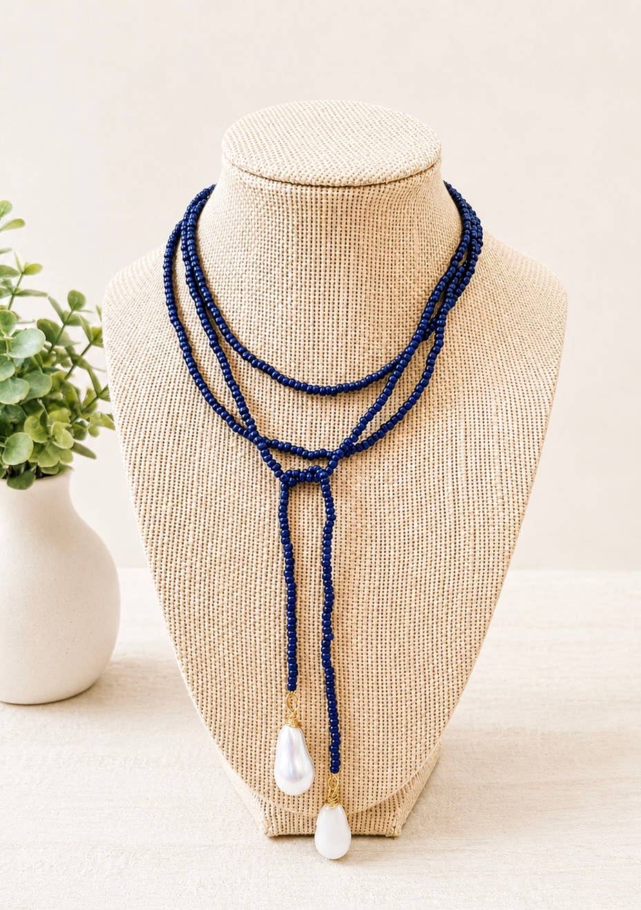 A multi-strand navy beaded lariat necklace with two white teardrop freshwater pearls, displayed on a beige woven jewelry bust.