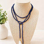 A multi-strand navy beaded lariat necklace with two white teardrop freshwater pearls, displayed on a beige woven jewelry bust.
