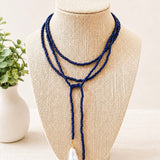 A multi-strand navy beaded lariat necklace with two white teardrop freshwater pearls, displayed on a beige woven jewelry bust.
