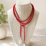 A red multi-strand beaded necklace with two white baroque pearl drop pendants, displayed on a beige fabric jewelry bust.