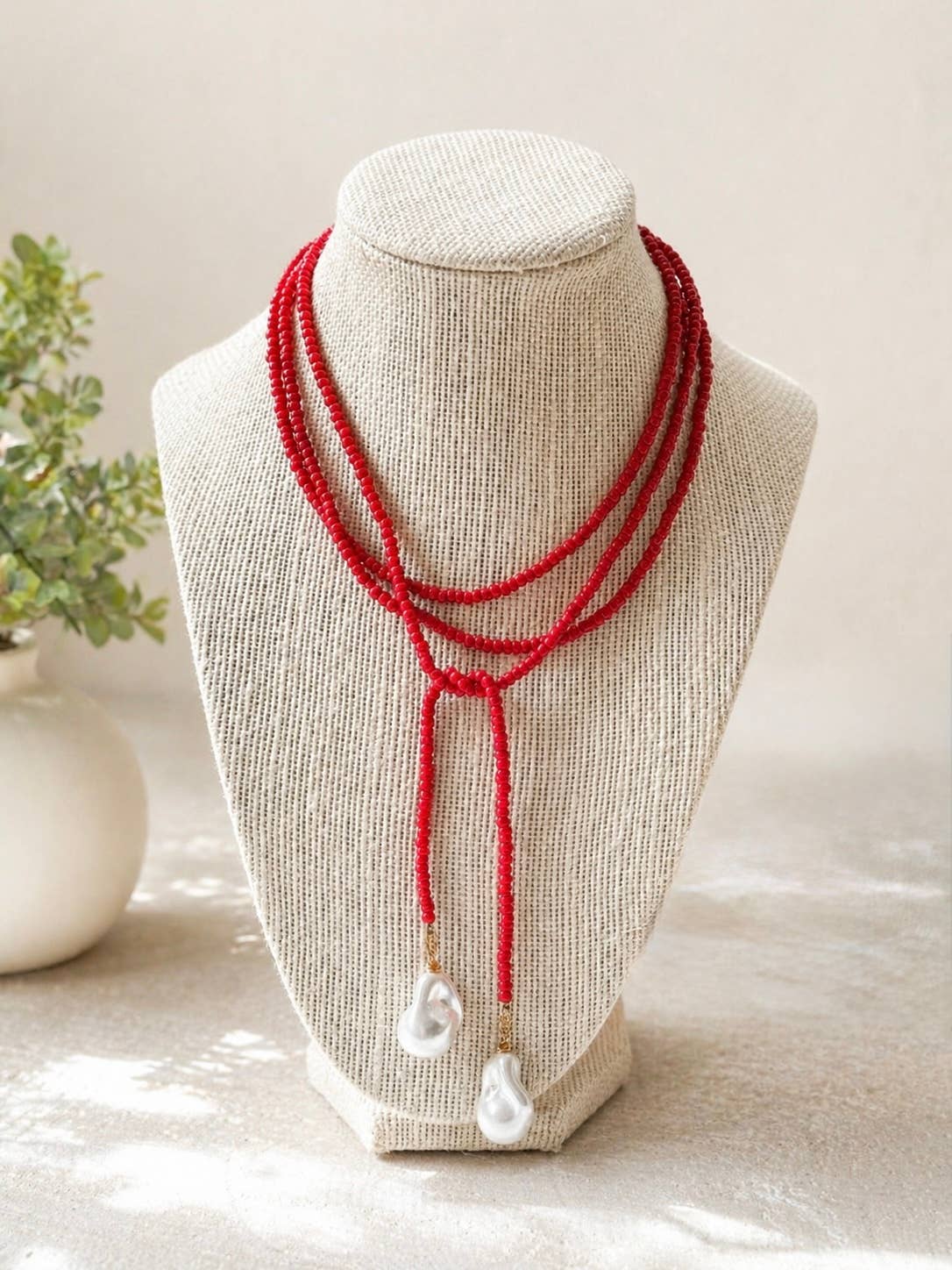 A red multi-strand beaded necklace with two white baroque pearl drop pendants, displayed on a beige fabric jewelry bust.