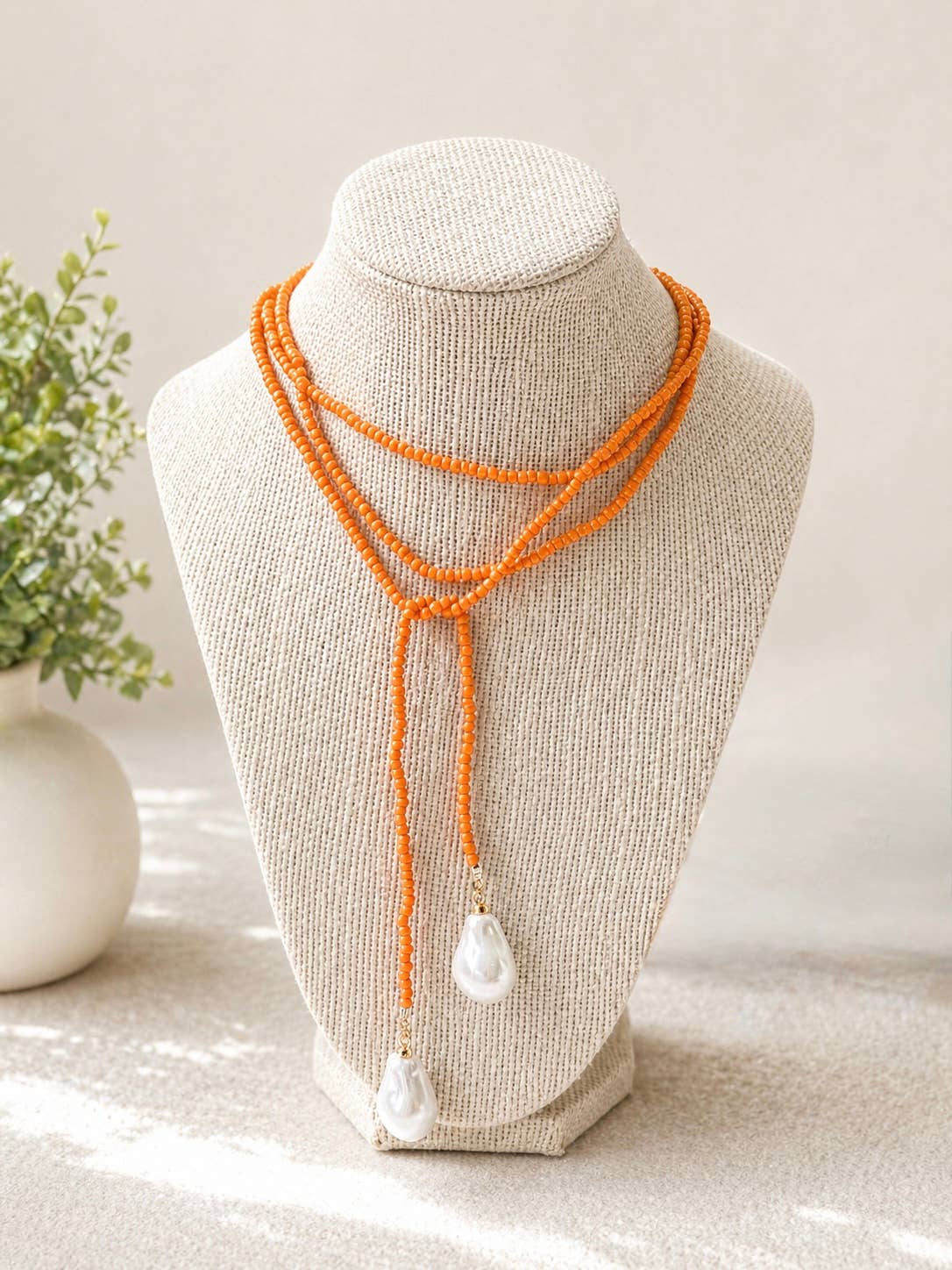 An orange beaded multi-strand necklace with two white teardrop pearl-like drops, displayed on a beige fabric jewelry bust.