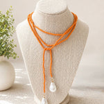 An orange beaded multi-strand necklace with two white teardrop pearl-like drops, displayed on a beige fabric jewelry bust.