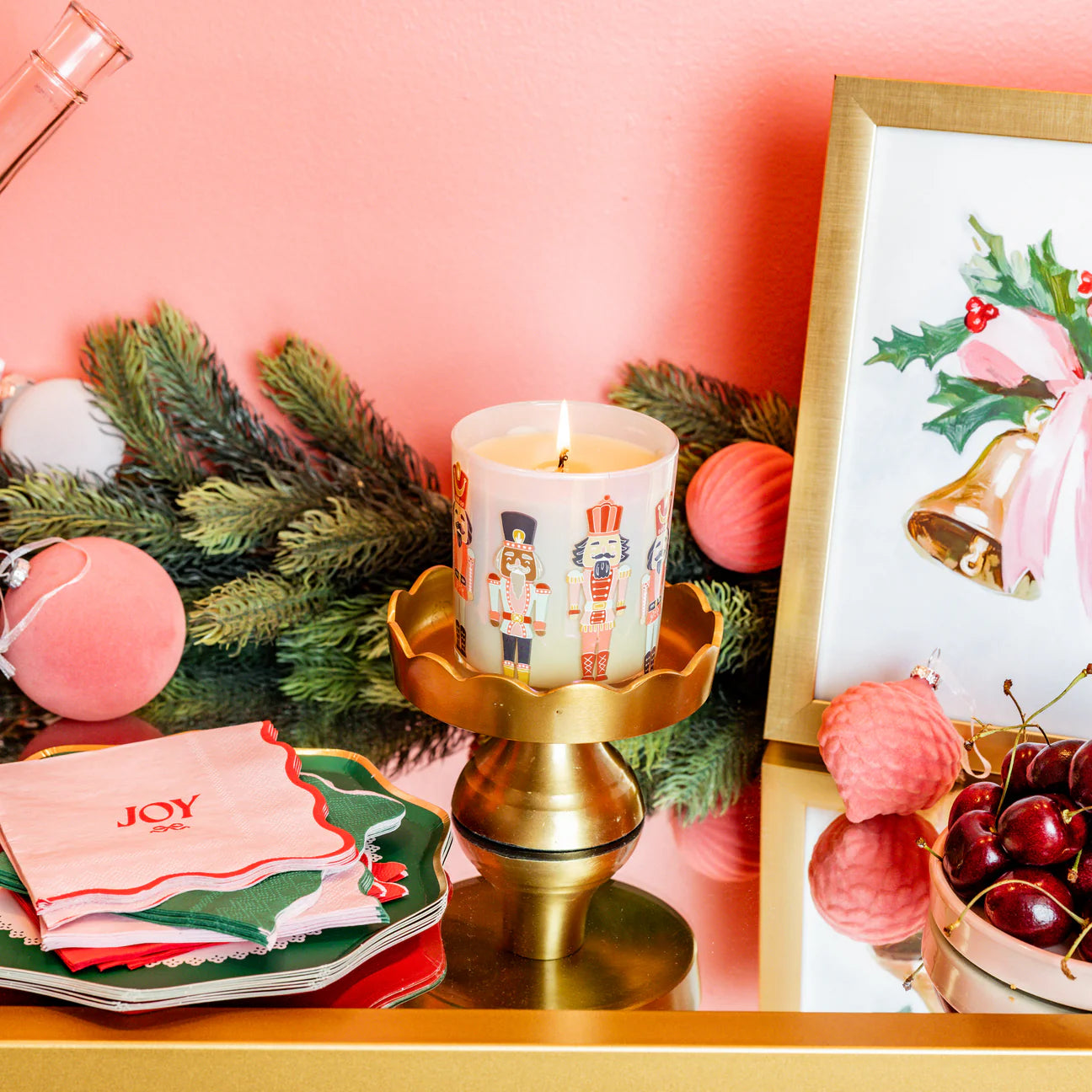 Decorative setup with a lit candle, Christmas-themed decor, and 'JOY' napkins on a pink background.