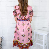 Woman wearing a pink dress with a pattern, standing against a white wooden panel wall.
