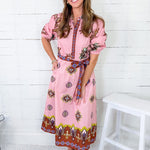 Woman wearing a pink dress with a pattern, standing against a white wooden panel background.