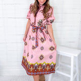 Woman wearing a pink patterned dress sitting on a white stool against a white wooden wall.