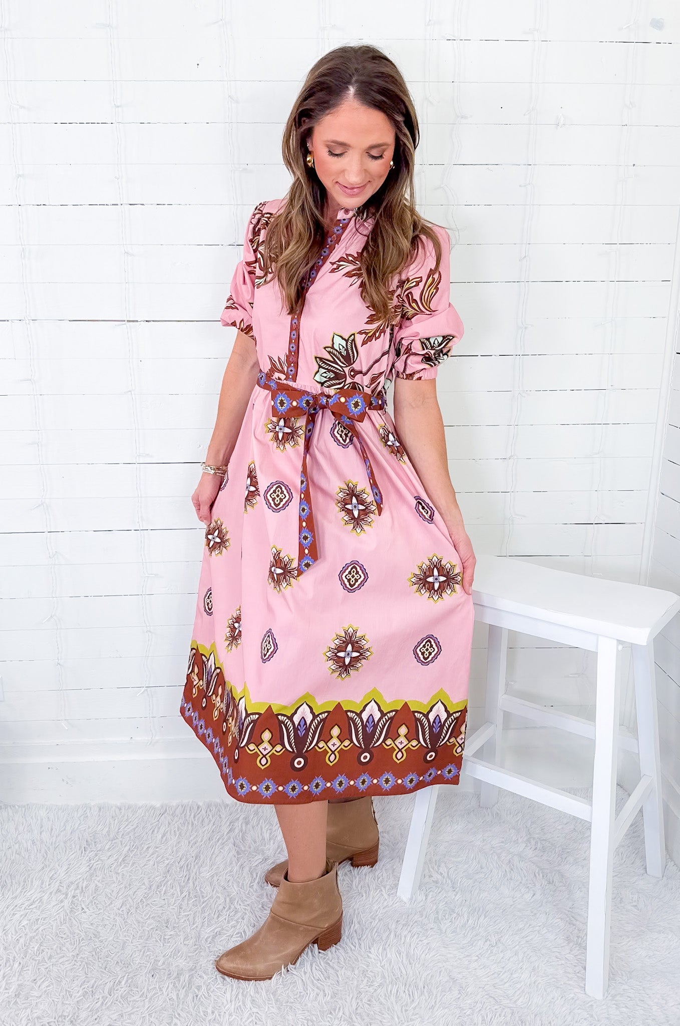 Woman wearing a pink patterned dress sitting on a white stool against a white wooden wall.