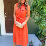 Woman in an orange dress standing on a porch with a brown door and greenery.