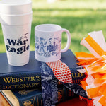 A styled outdoor product display featuring a white foam cup and a white ceramic mug with blue graphics, placed on top of a navy blue Webster's Dictionary book. A navy blue bandana with a red-and-white check pattern is tied around the book, and orange-and-white pom-poms sit to the right.