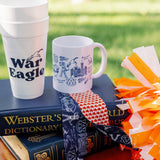 A styled outdoor product display featuring a white foam cup and a white ceramic mug with blue graphics, placed on top of a navy blue Webster's Dictionary book. A navy blue bandana with a red-and-white check pattern is tied around the book, and orange-and-white pom-poms sit to the right.