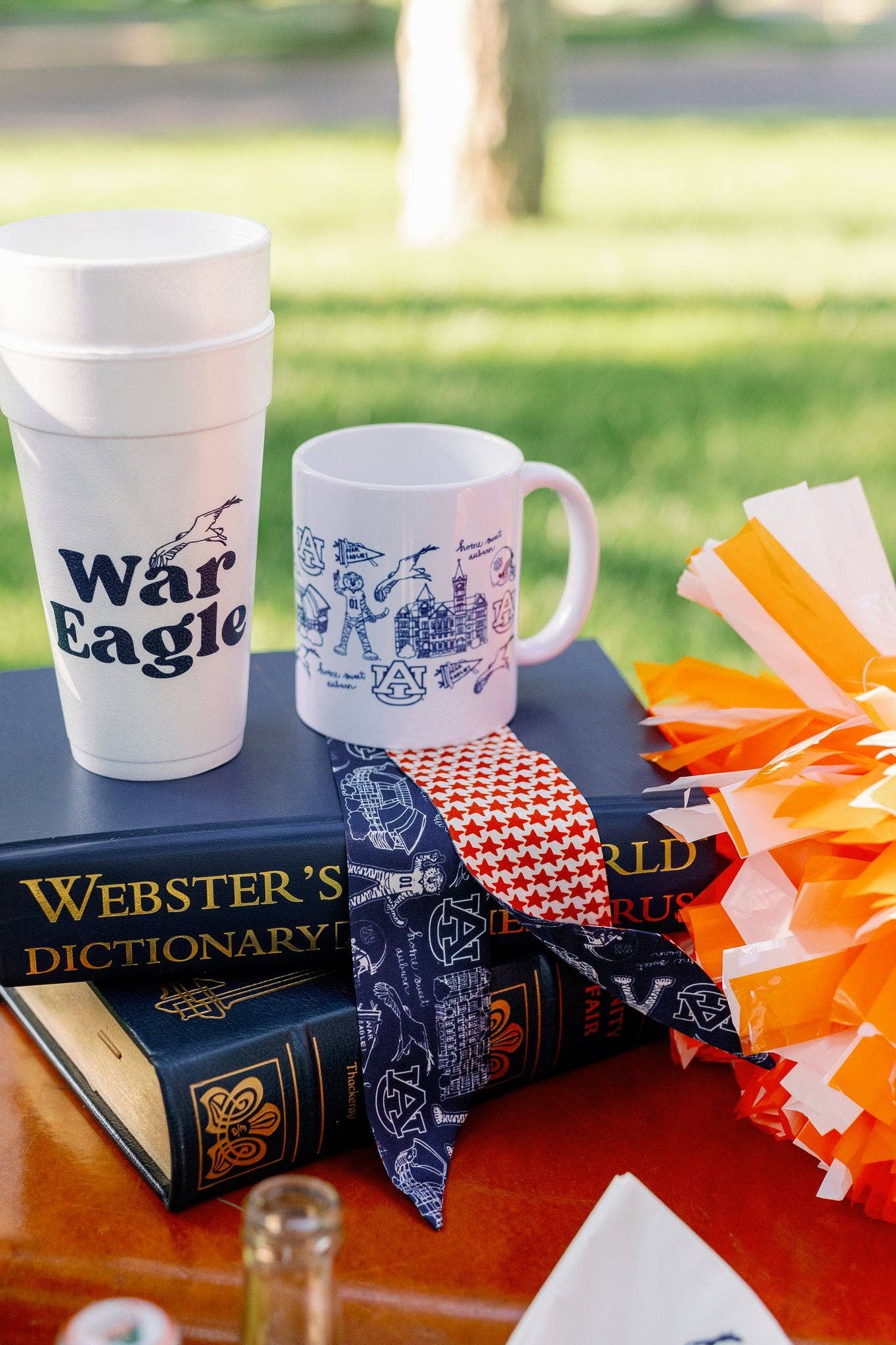 A styled outdoor product display featuring a white foam cup and a white ceramic mug with blue graphics, placed on top of a navy blue Webster's Dictionary book. A navy blue bandana with a red-and-white check pattern is tied around the book, and orange-and-white pom-poms sit to the right.