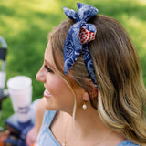 A woman outdoors wearing a blue patterned fabric headband with a red and white polka dot accent, paired with a light blue denim top.