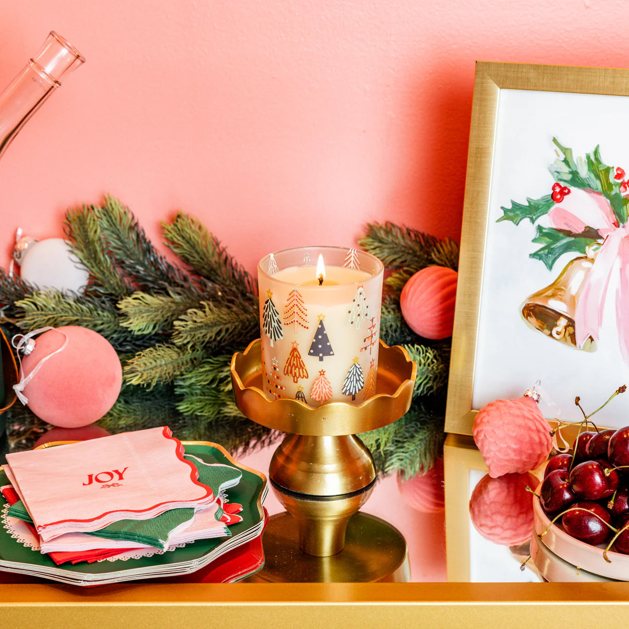 Decorative table setting with a lit candle, Christmas-themed decor, and a pink background.